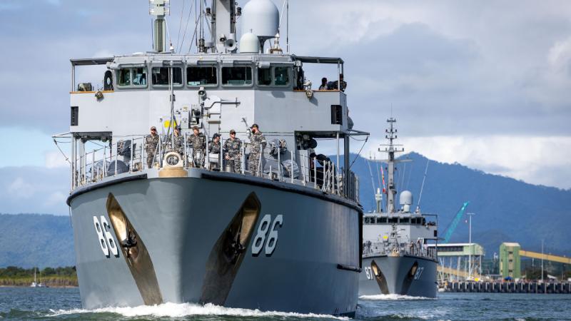 Two naval ships sailing out of port, with people visible on the deck of the leading ship.