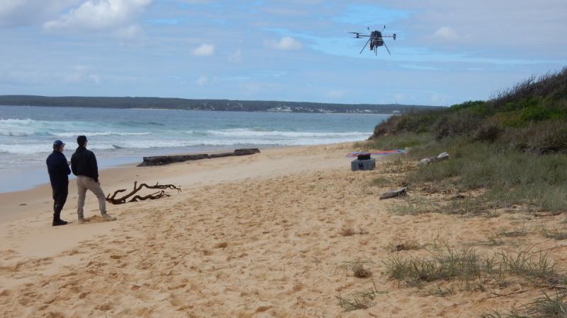 Two people stand on a beach as a drone flies overhead.
