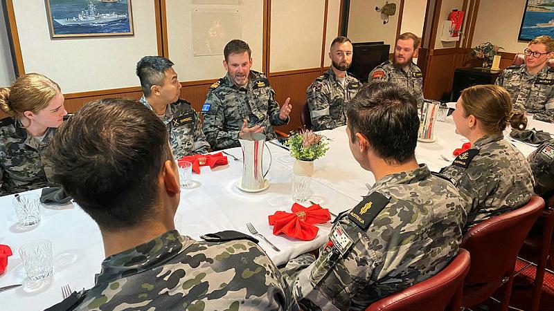 A group of Navy personnel sit at a dining table as they are addressed by one individual.