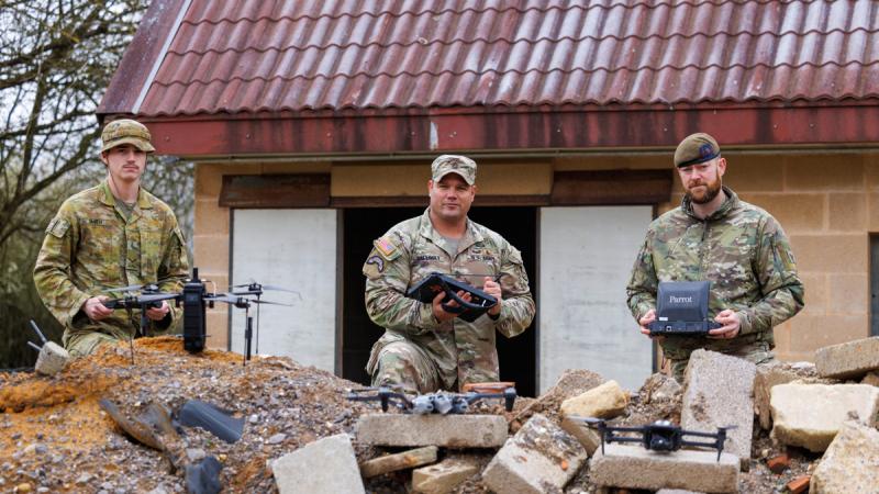Three drones rest on a pile of rubble in front of three men in camouflage uniforms holding devices in their hands.