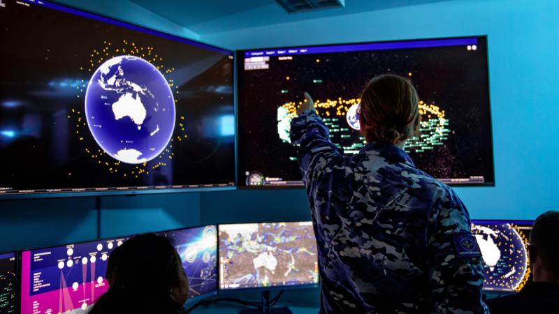 A woman in camouflage uniform points at maps on wall-mounted screens.