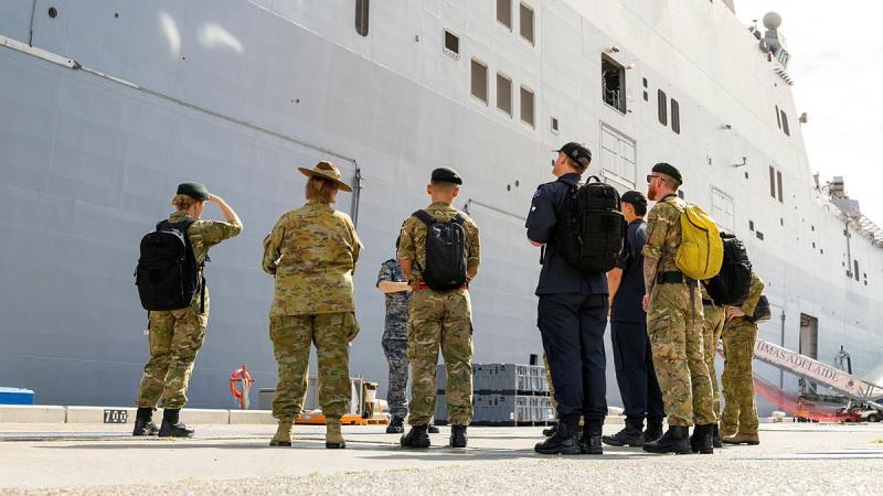 A group of New Zealand Defence personnel stand and look up at a ship from a dock.