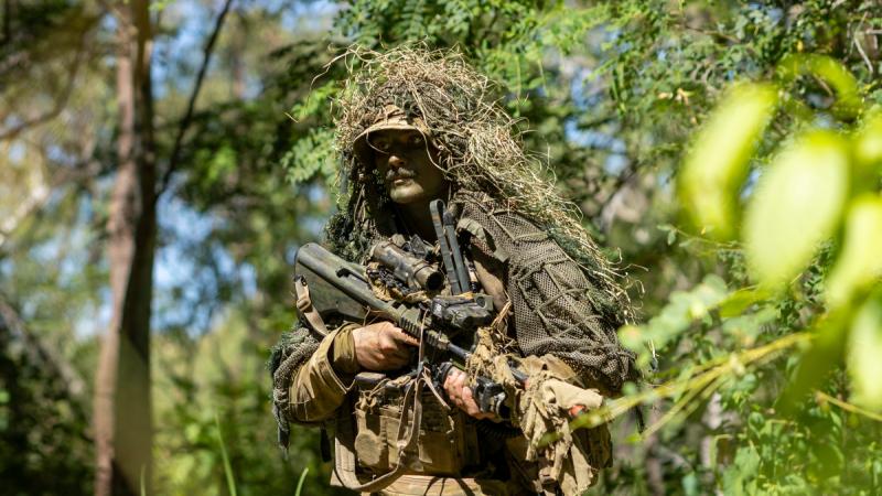 A man in camouflage and a ghillie suit holds a camouflaged firearm in dense vegetation.