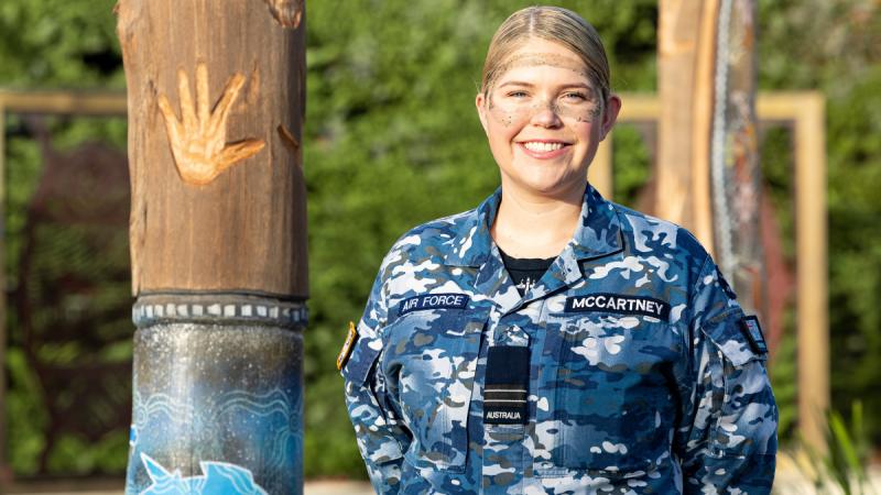 A woman in Air Force camouflage and ochre face paint around her eyes stands next to some large wooden poles decorated with Indigenous artwork.