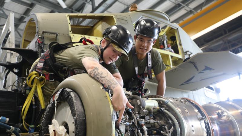Two Army personnel lean over and work on parts of a helicopter.