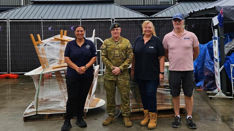 Four people, one in uniform, stand in a row with furniture and other items wrapped in plastic on pallets behind them.