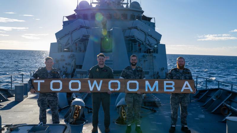 Four men in uniform stand in line on a ship's deck holding a long wooden sign with large silver letters spelling ‘TOOWOOMBA’.