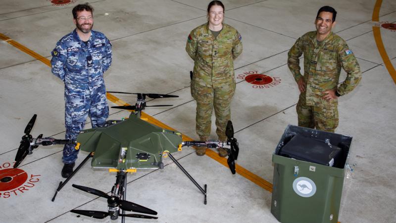 Three people in uniform standing behind a green drone and a large green box.