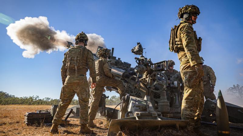 People in uniform stand beside an artillery gun as smoke billows from its barrel.