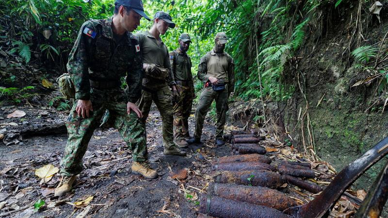 Four military personnel stand facing a row of unexploded ordnance on the ground in a jungle setting.