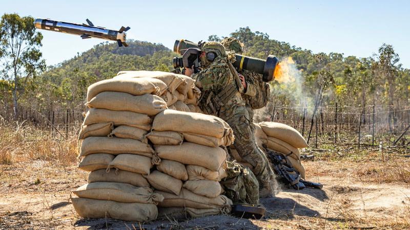 A soldier leans on sandbags and fires a large weapon, the projectile is seen in the air with flames at the back of the weapon.