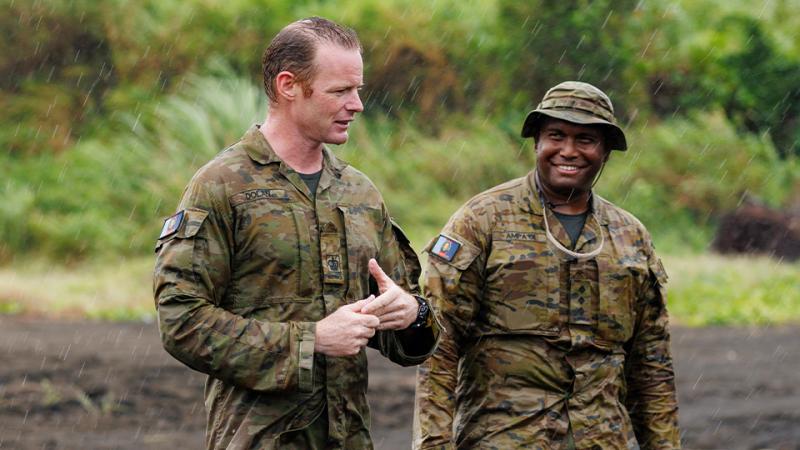 Two men in uniform stand outdoors in rainy conditions with dense vegetation behind them.