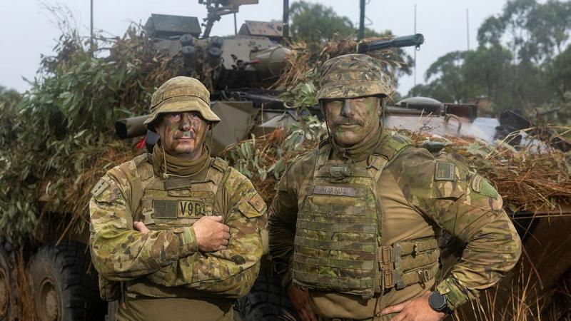 Two soldiers stand together in front of a camouflaged Army vehicle.