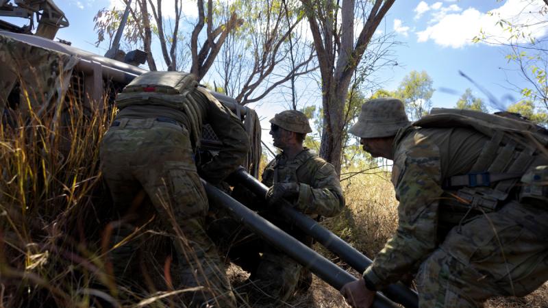 Three men in camouflage uniform attach large metal poles to a vehicle.
