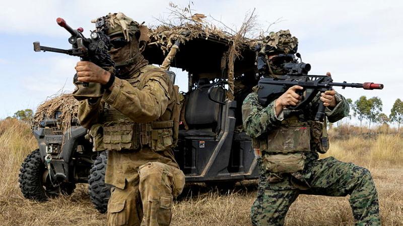 Two soldiers hold and aim guns in front of an Army vehicle in a grassy setting.