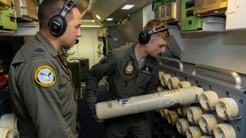 A man in a flight uniform inserts a large cylindrical object into a rack inside an aircraft cabin while another man watches.