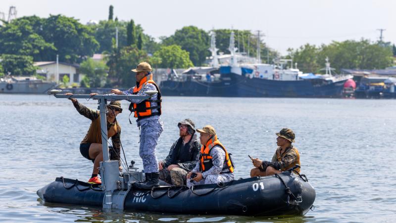 Five people in a small inflatable boat beside a large T-shaped metal frame.