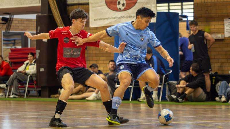 A man in a blue sports shirt kicking a soccer ball on an indoor court as a man in a red sports shirt tries to block him.