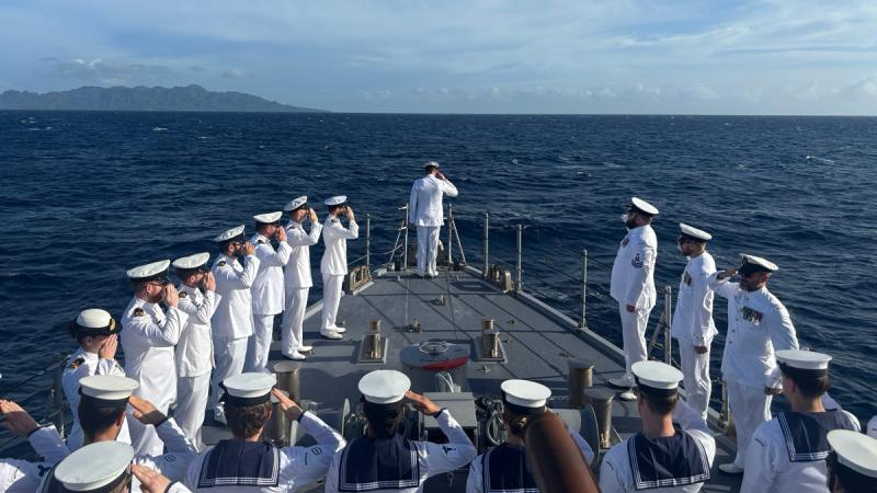 People in Navy uniforms salute at the bow of a ship, with land visible on the horizon.