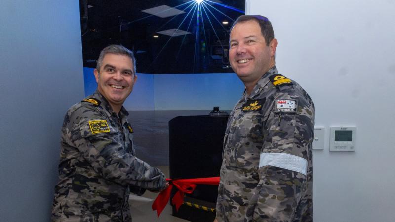 Two men in uniform stand beside an open doorway as one cuts a red ribbon.