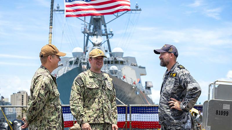 Three Navy personnel from Australia and the US stand and talk. A ship is seen in the background.