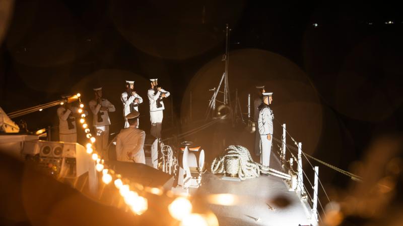 Several uniformed sailors stand at the front of a ship at night, with some carrying rifles.