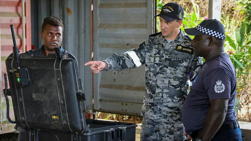 Two islander men in police uniforms talk to an Australian Navy member about equipment in a hard case in front of them.