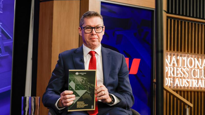 A man in a suit sitting down while holding a booklet up.