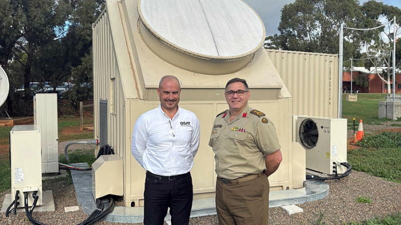 Two men stand in front of a small structure in an open bush area.