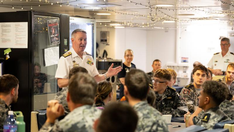 A man in a white uniform speaks to seated people in camouflage uniforms in a room with vending machines.