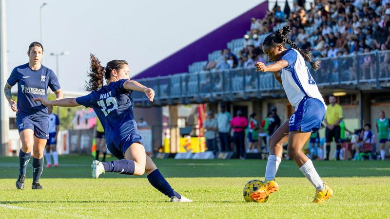 A soccer player runs on an angle and prepares to kick a ball as she faces opposition.