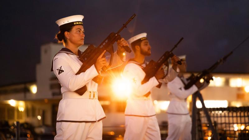 Three people in Navy uniforms hold rifles angled upwards at night.