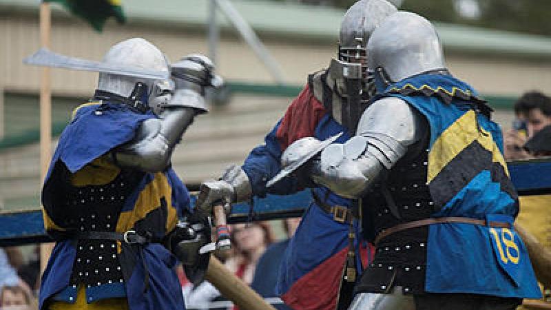 Three people dressed in metal armour engage in sporting combat with swords.