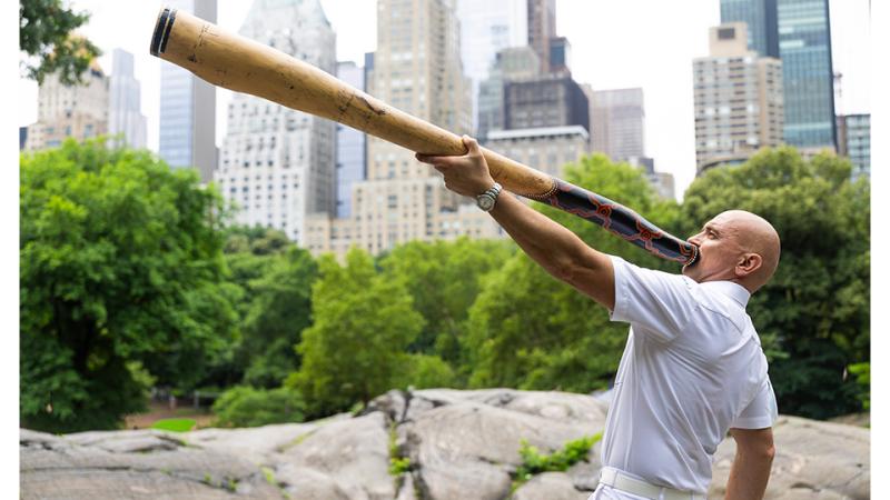 A man all in white plays a didgeridoo in a city park.