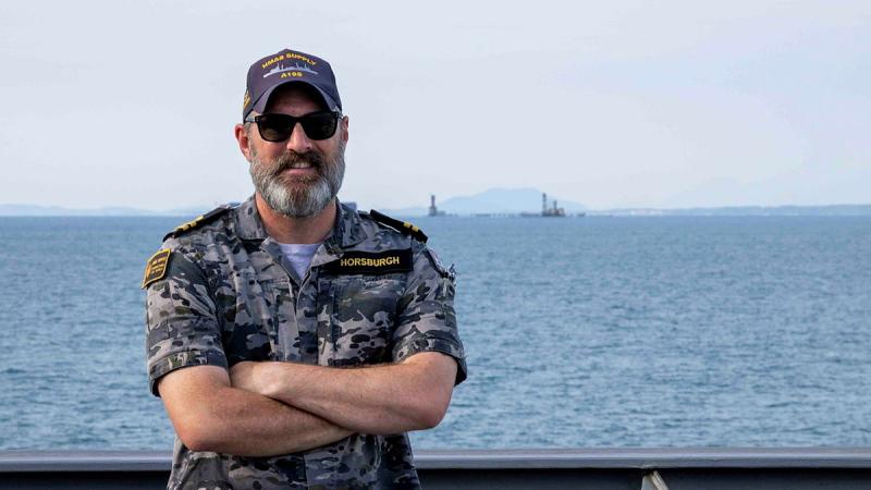 A Navy member stands on the deck of a ship with his arms crossed. The ocean and a light house are seen in the background.