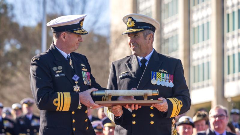 A male Navy officer in dress uniform hands the weight of command to a fellow officer in front of a crowd outside.
