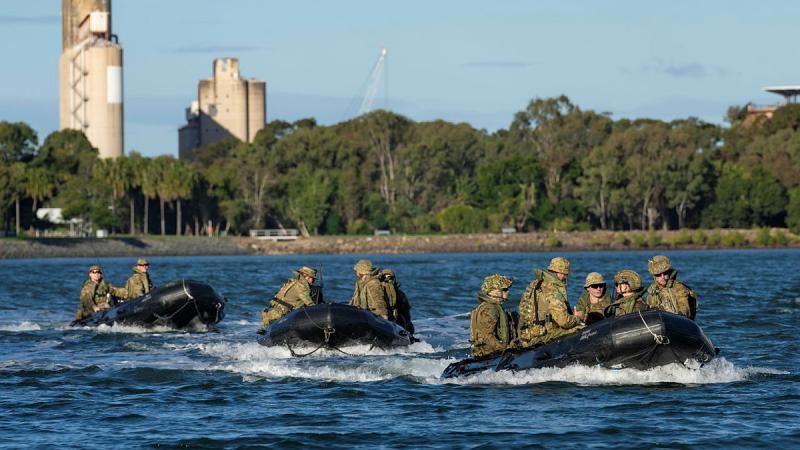 Soldiers in small watercraft move across a body of water with buildings and vegetation in the background.