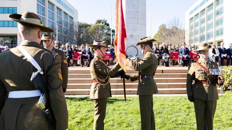 A male and female Army officer exchange a red flag in front of a memorial stand of seated spectators.