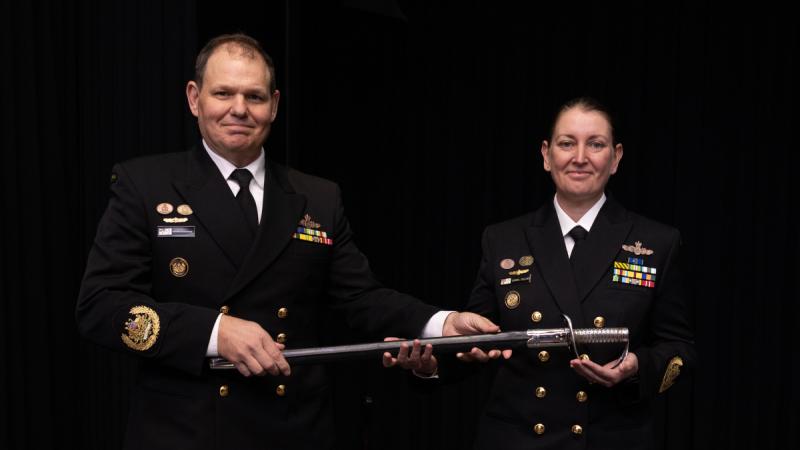 A male Navy officer hands a ceremonial sword to a female Navy officer on a theatrette stage.