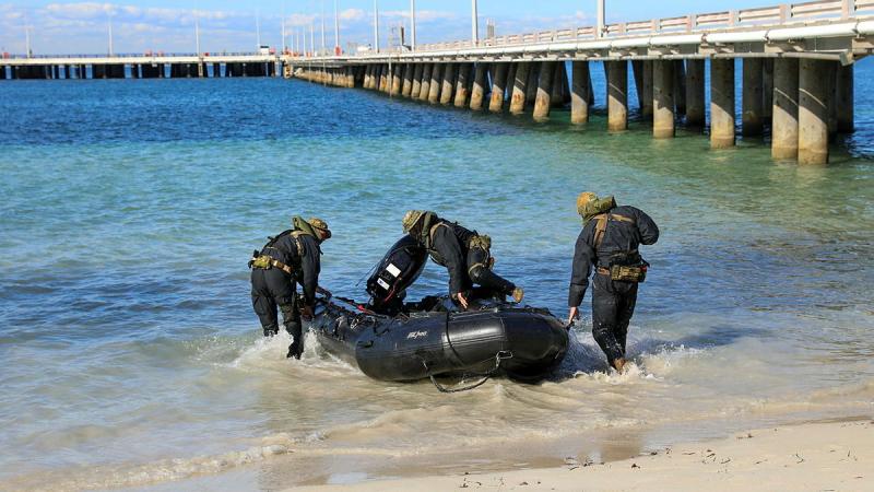 A few people hold a small black watercraft and walk into water near a boardwalk.