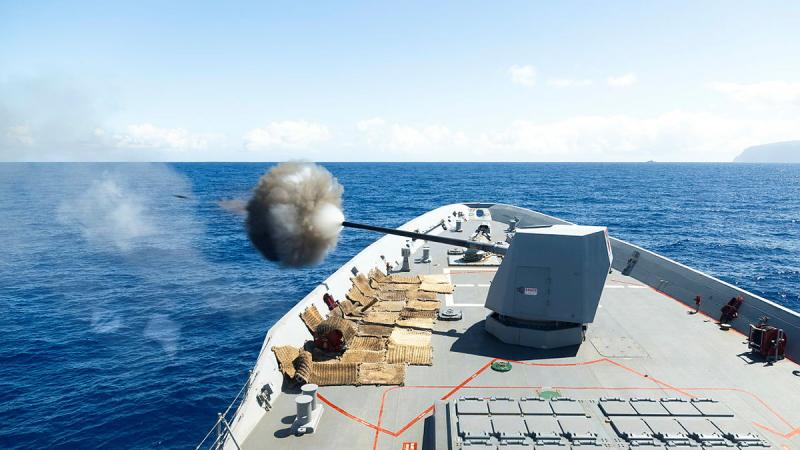 A large gun on a ship fires out to sea with a dark cloud of smoke seen and a projectile.