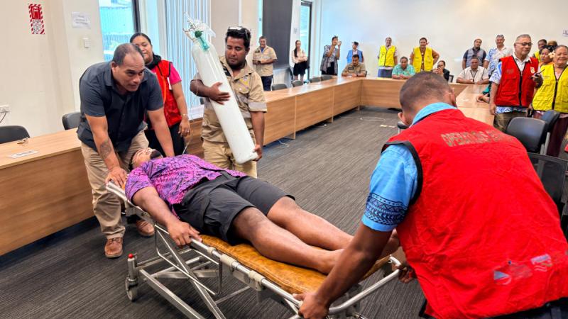 Two people move a man on a stretcher while another stands nearby holding a large cylindrical breathing apparatus.