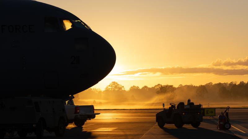 A large aircraft is silhouetted against a setting sun on a tarmac with a small vehicle near it.