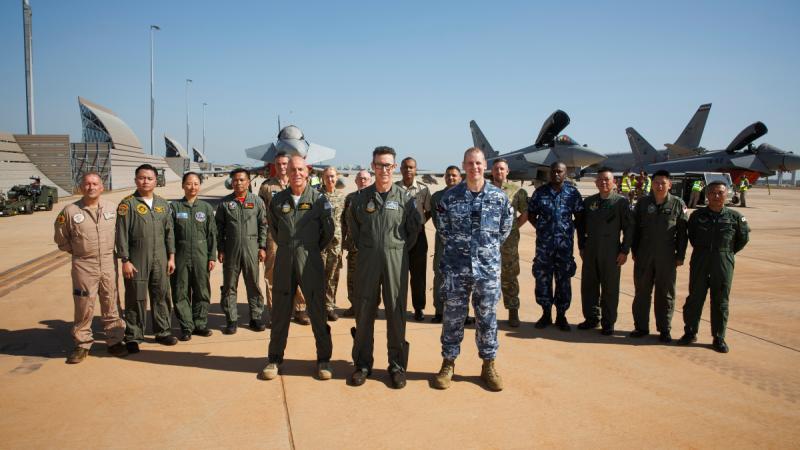 A group of military personnel standing together on the tarmac is aircraft in the background.