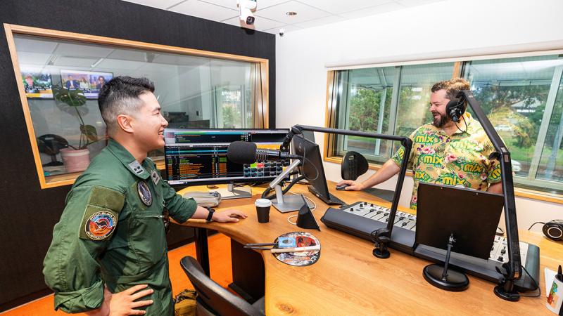 A person in a green flight suit stands beside a desk in a radio broadcasting studio, speaking with a headset-wearing presenter across a microphone-equipped broadcast console. Multiple computer monitors, microphones on adjustable boom arms, and audio control equipment are visible on the desk, while large windows look into adjacent rooms and outside greenery.
