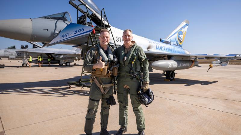 Two pilots stand together, smiling, in front of two aircraft on a tarmac.