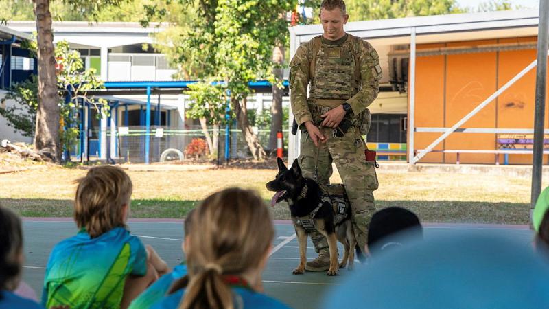 A military man stand with a dog on a lead as children watch.