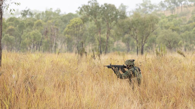 A soldier aims his gun as he moves through long grass.