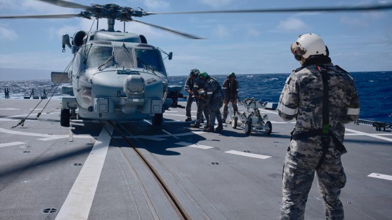 People in uniforms and helmets lifting a missile onto a military helicopter on a ship’s flight deck.