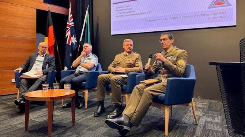 A civilian and three military men sit for a discussion panel with a PowerPoint presentation.